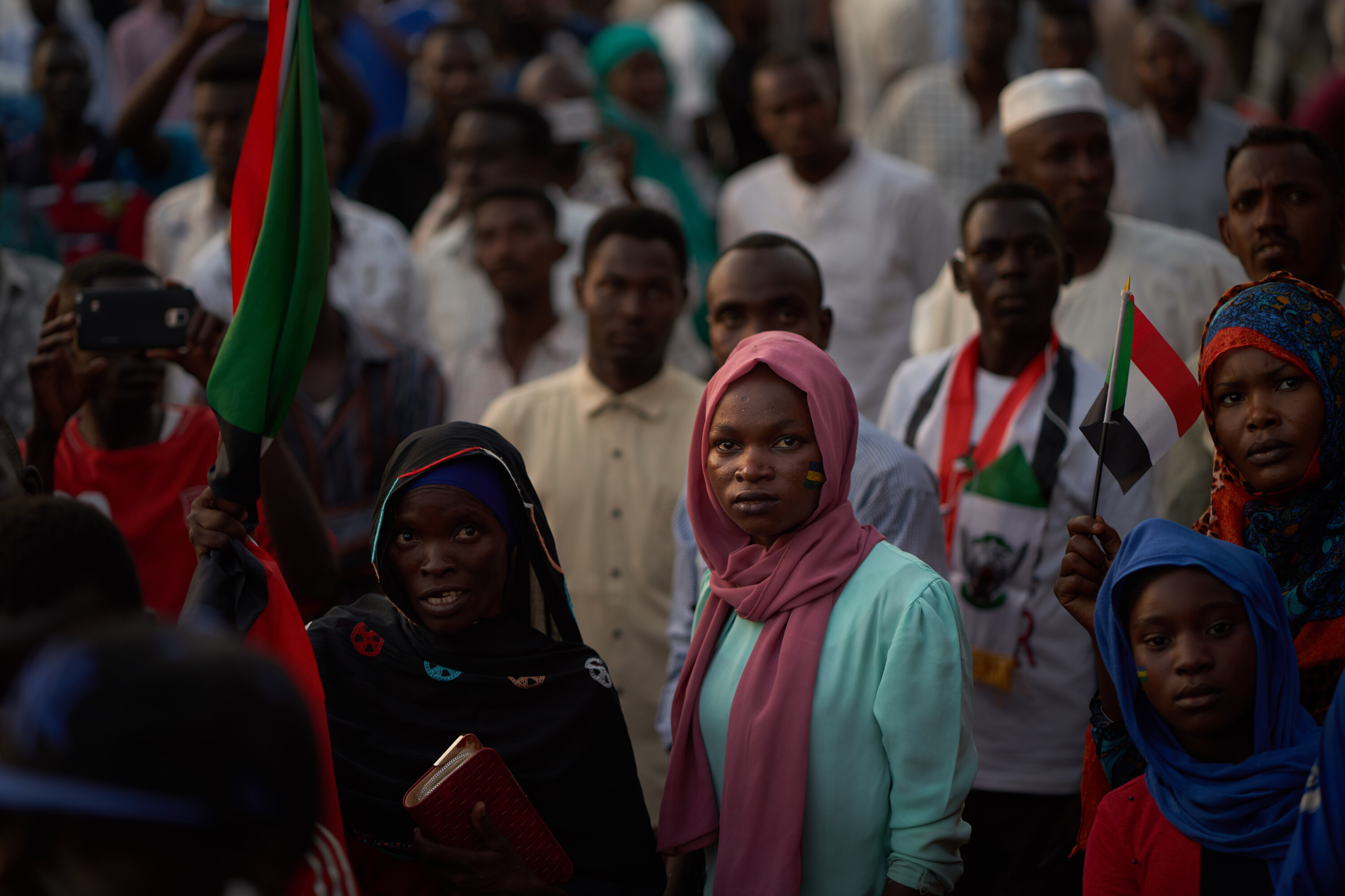 Women with Sudanese flags stand at the front of a crowd; a woman in a pink headscarf and mint shirt faces forward.