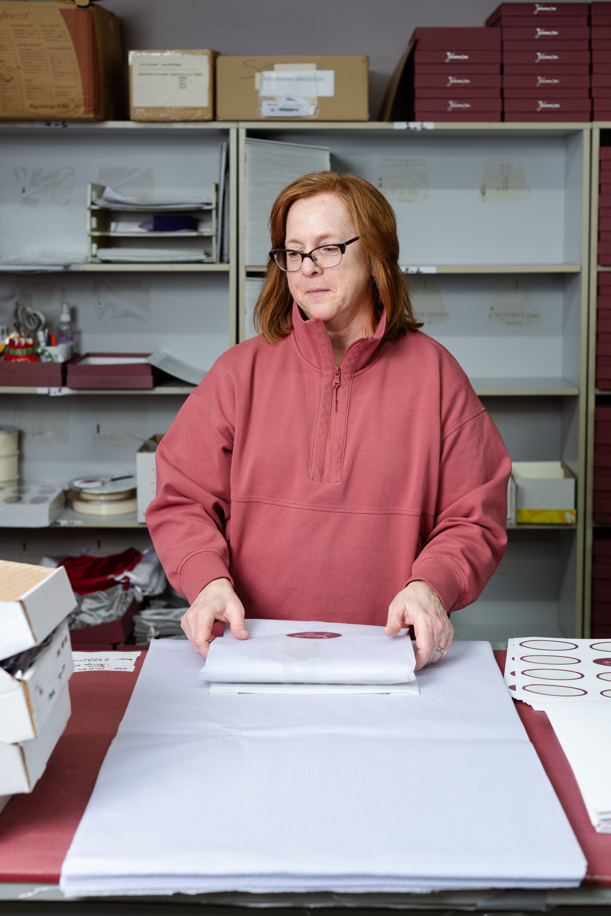 A worker in a pink pullover folds white tissue around a garment at a packing table.